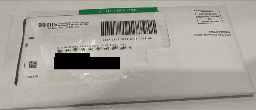 A certified mail envelope from the IRS lying on a kitchen table, name and address blacked out.